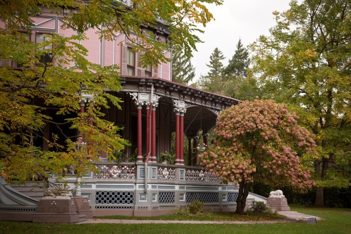 A covered porch with red columns and a white and gray railing wraps around a pink house