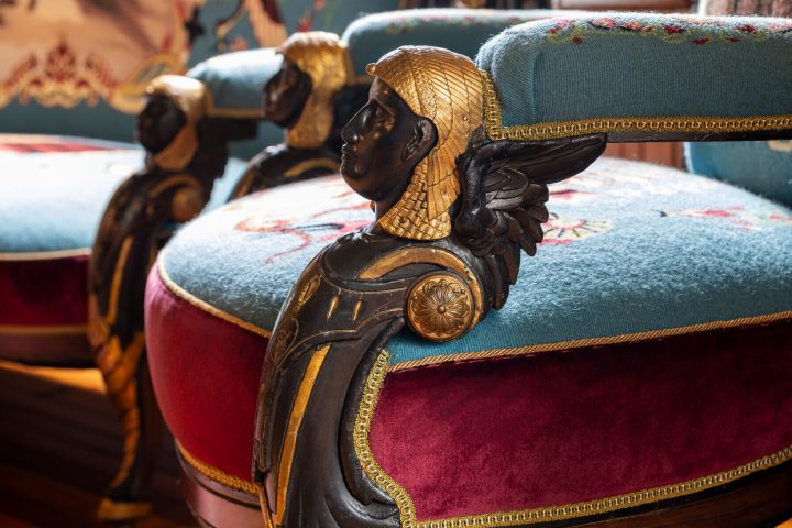 The arm of a chair carved to look like Cleopatra, with a wood face and bronze hair.