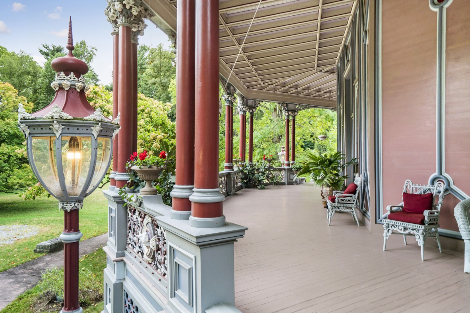 Victorian-style porch with ornate columns, plants, and chairs with red cushions.
