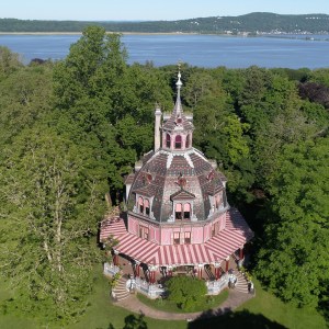 Circular Victorian house with red roof surrounded by trees near a large body of water.