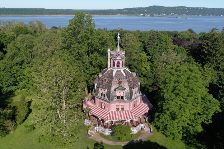 Circular Victorian house with red roof surrounded by trees near a large body of water.