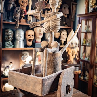 Skeleton statue in a wooden cart, surrounded by masks on shelves.